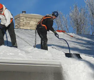 Professional technicians performing roof snow removal with safety harnesses and shovels in Arvada, CO